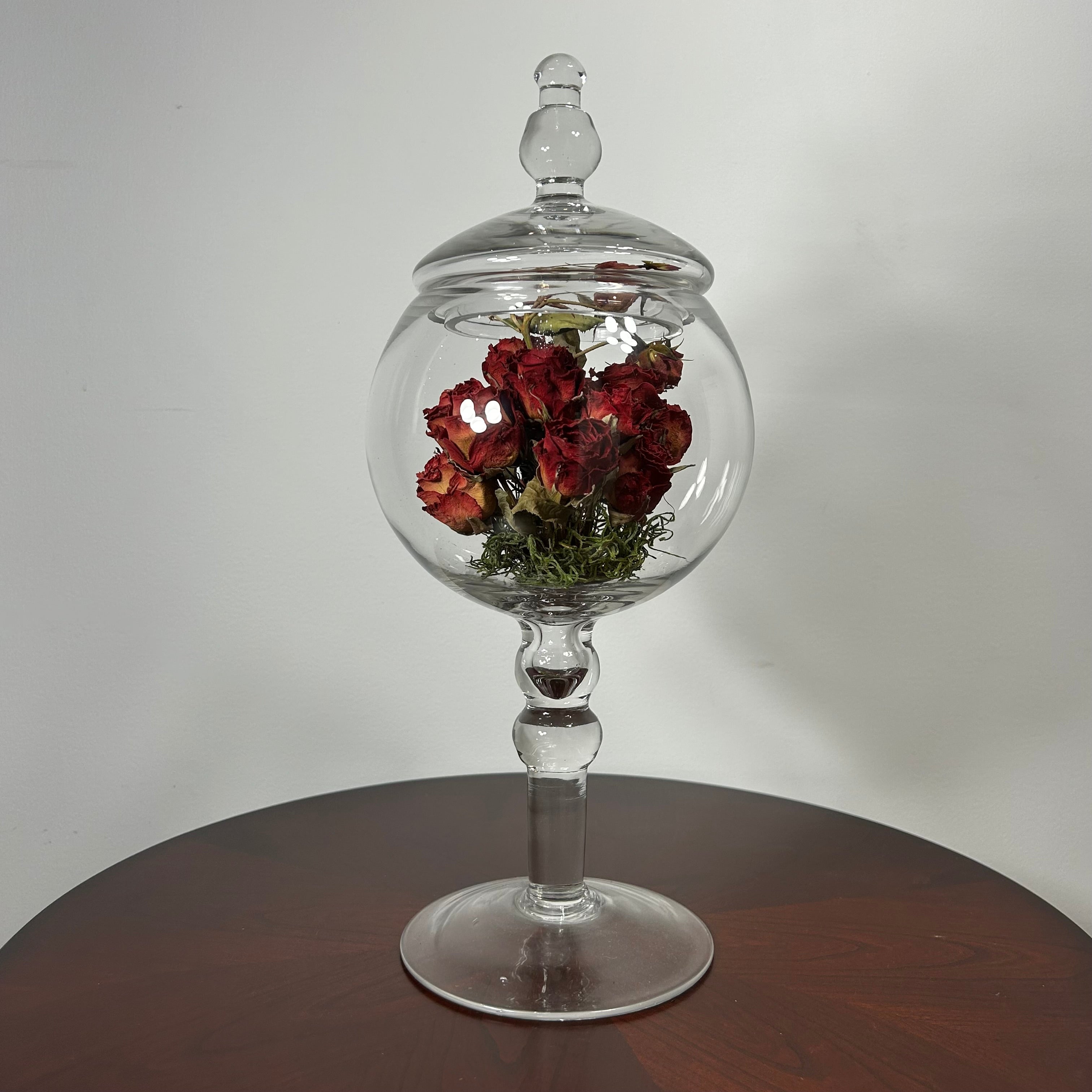 A clear glass apothecary-style jar with a lid and pedestal base, displayed on a round wooden table against a light gray wall. Inside the glass dome, dried red roses with green stems are arranged.