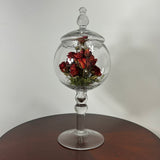 A clear glass apothecary-style jar with a lid and pedestal base, displayed on a dark wooden table against a plain light wall. Inside the glass dome are dried red roses with green stems.
