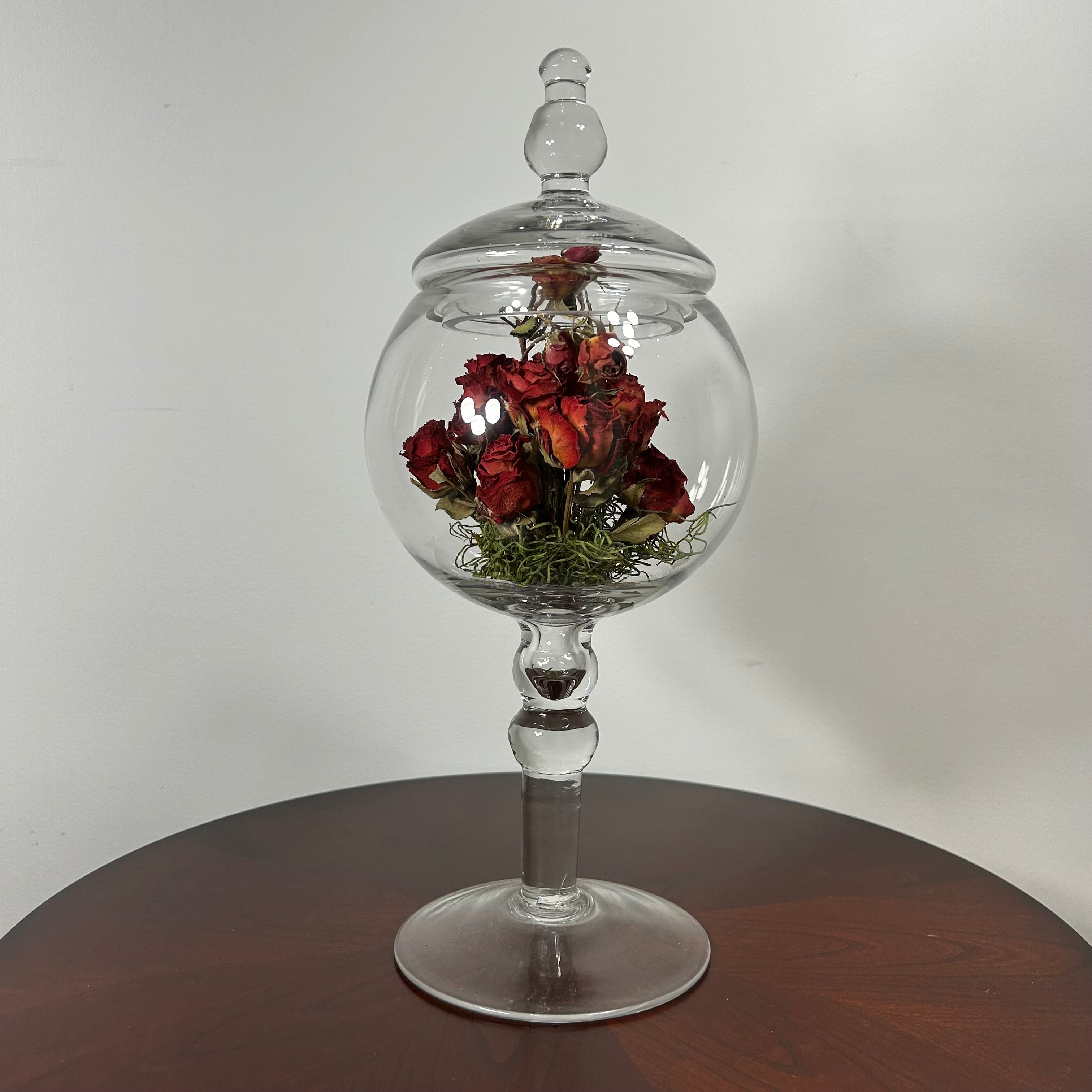 A clear glass apothecary-style jar with a lid and pedestal base, displayed on a dark wooden table against a plain light wall. Inside the glass dome are dried red roses with green stems.