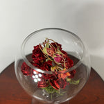 A clear glass fishbowl vase filled with dried red roses and green stems, placed on a dark wooden surface against a plain light gray wall.