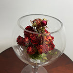 A bouquet of dried red roses arranged inside a clear glass goblet or bowl-shaped vase, set on a dark wooden surface against a plain light background.