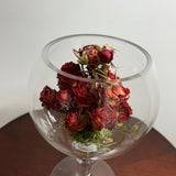 A bouquet of dried red roses arranged inside a clear glass goblet or bowl-shaped vase, set on a dark wooden surface against a plain light background.