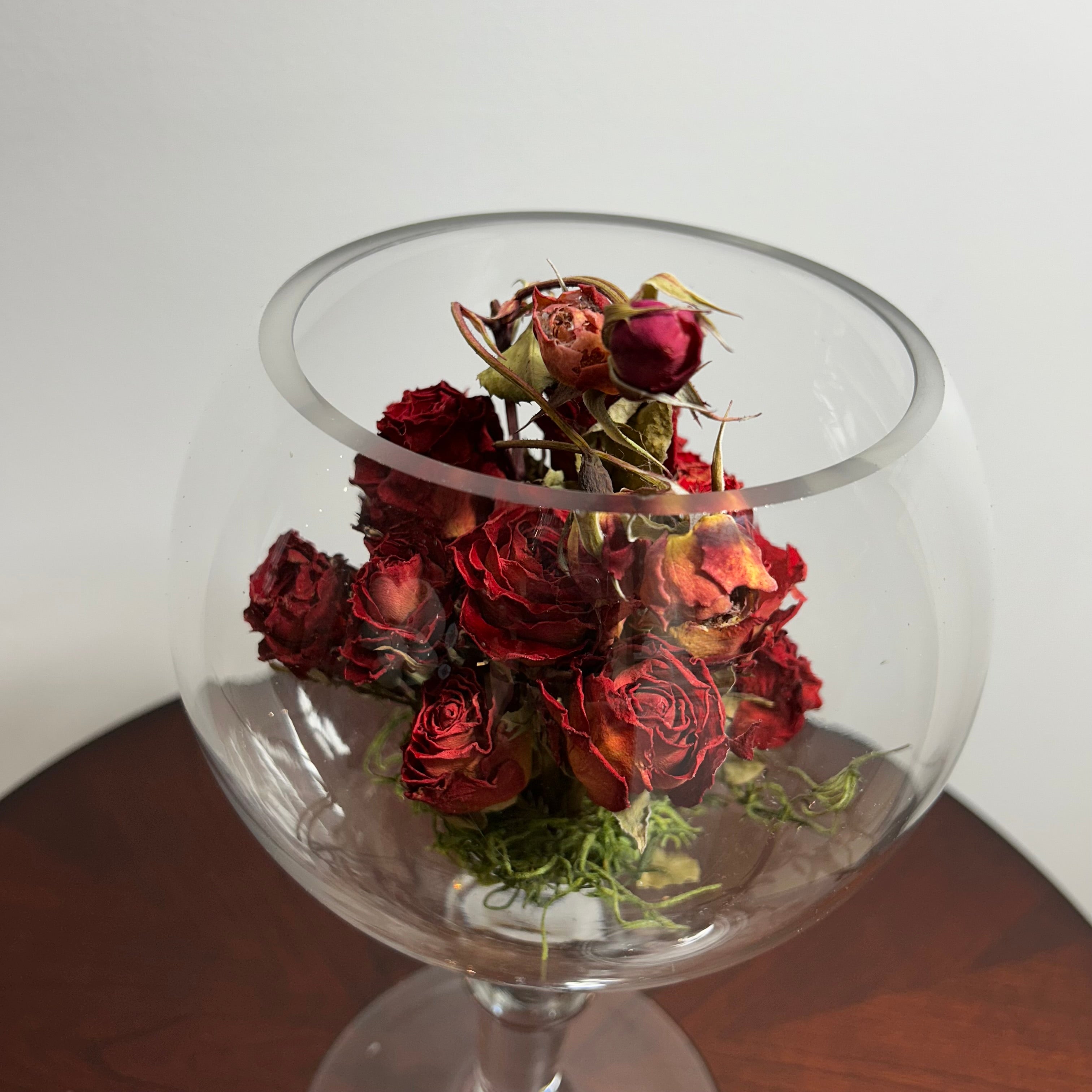 A bouquet of dried red roses arranged inside a clear glass goblet or bowl-shaped vase, set on a dark wooden surface against a plain light background.