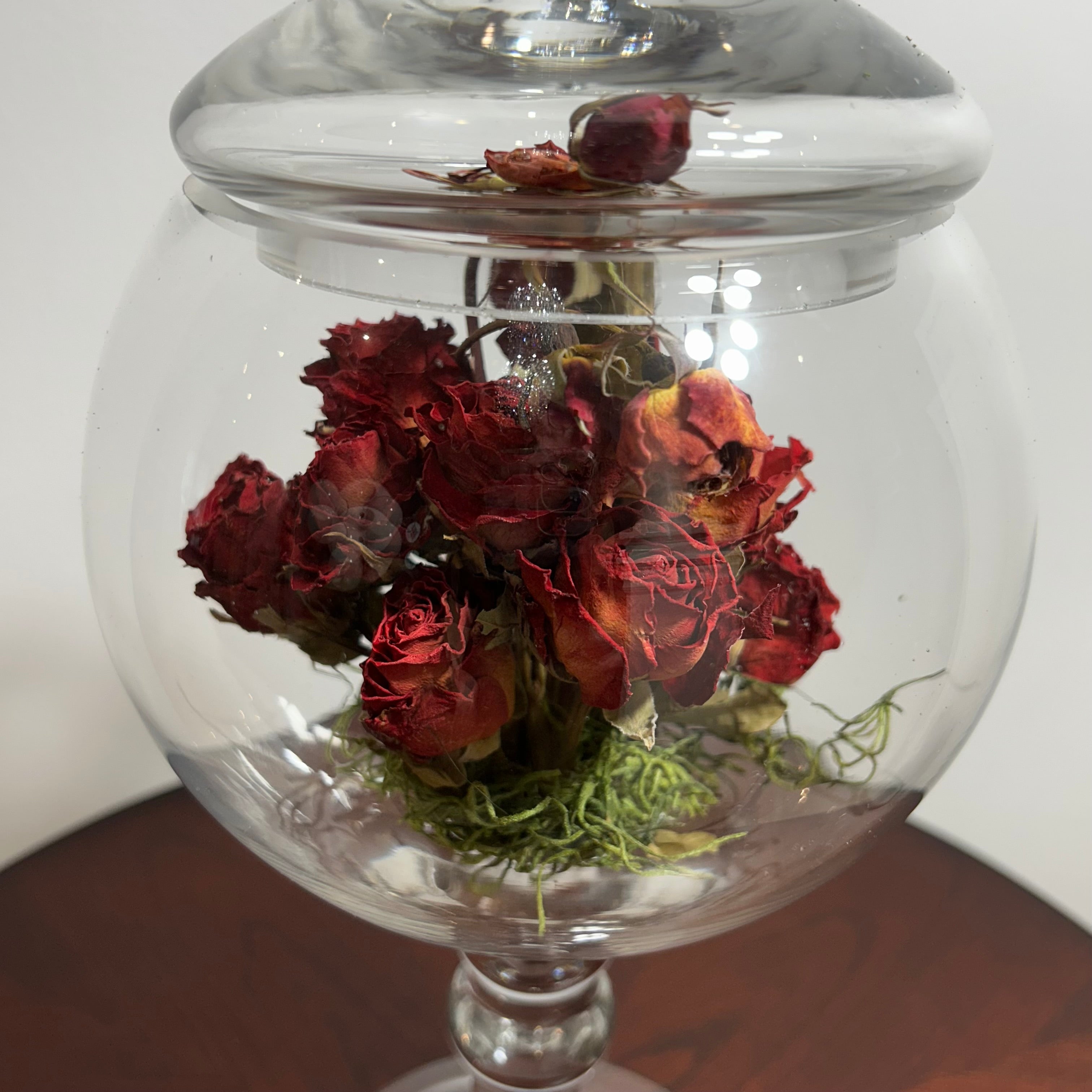 A clear glass jar with a lid, filled with dried red roses and green moss, set on a dark wooden surface against a plain light background.