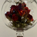 A clear glass fishbowl-style vase with a pedestal base holding a bouquet of dried red roses with green moss at the bottom, photographed against a plain light wall.