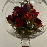 A clear glass fishbowl-style vase with a pedestal base holding a bouquet of dried red roses with green moss at the bottom, photographed against a plain light wall.