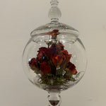 A clear glass terrarium-style jar with a rounded body, lid, and pedestal base, containing dried red roses and green moss against a plain light wall.
