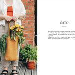 Person holding a yellow tote bag with plants, standing next to a brick wall.