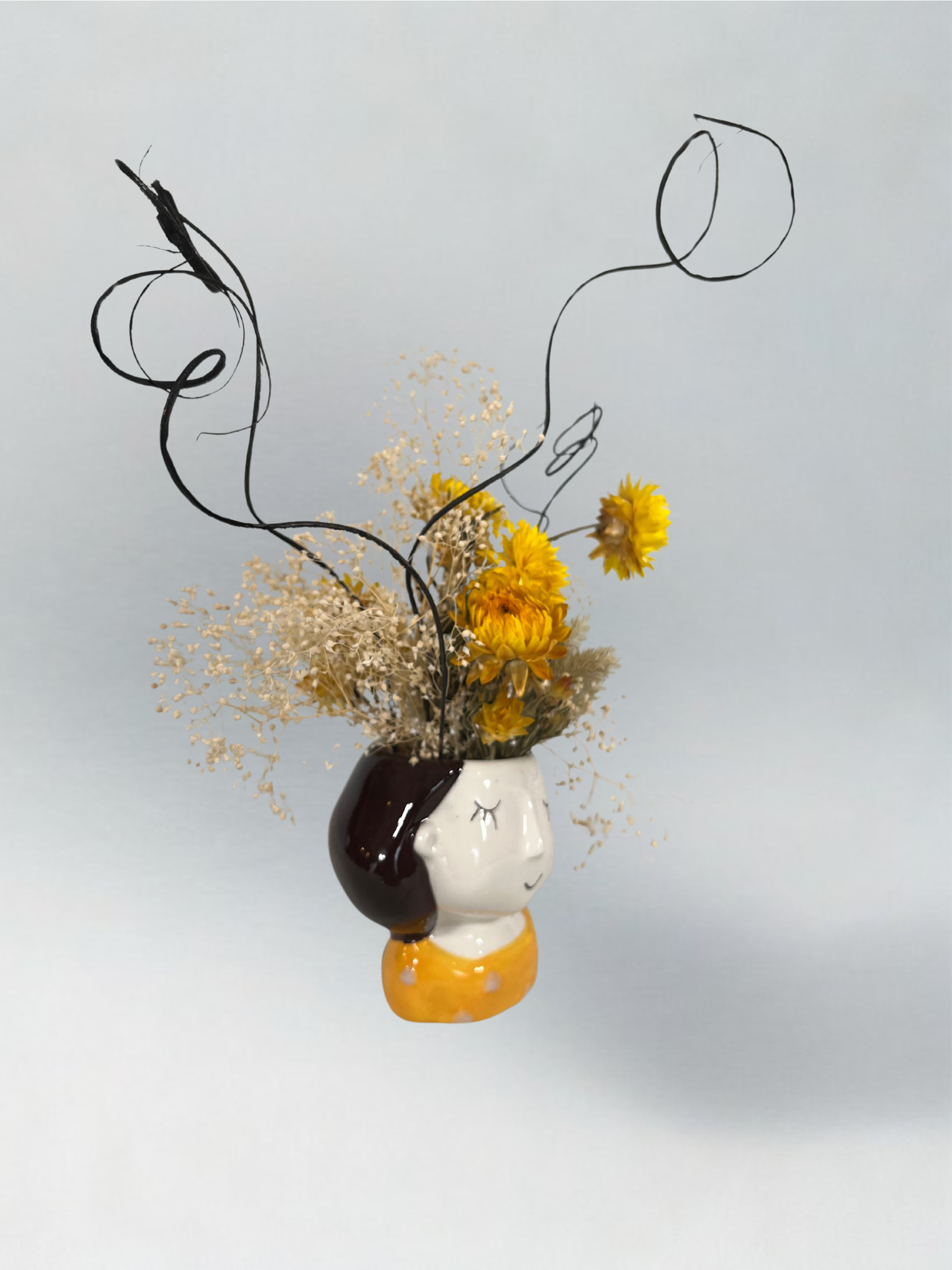 Decorative head-shaped vase with yellow flowers and brown curly ting ting accents on a light grey background