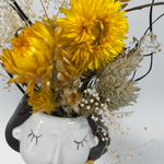 Vase shaped like a face with yellow flowers and dried elements on a white background