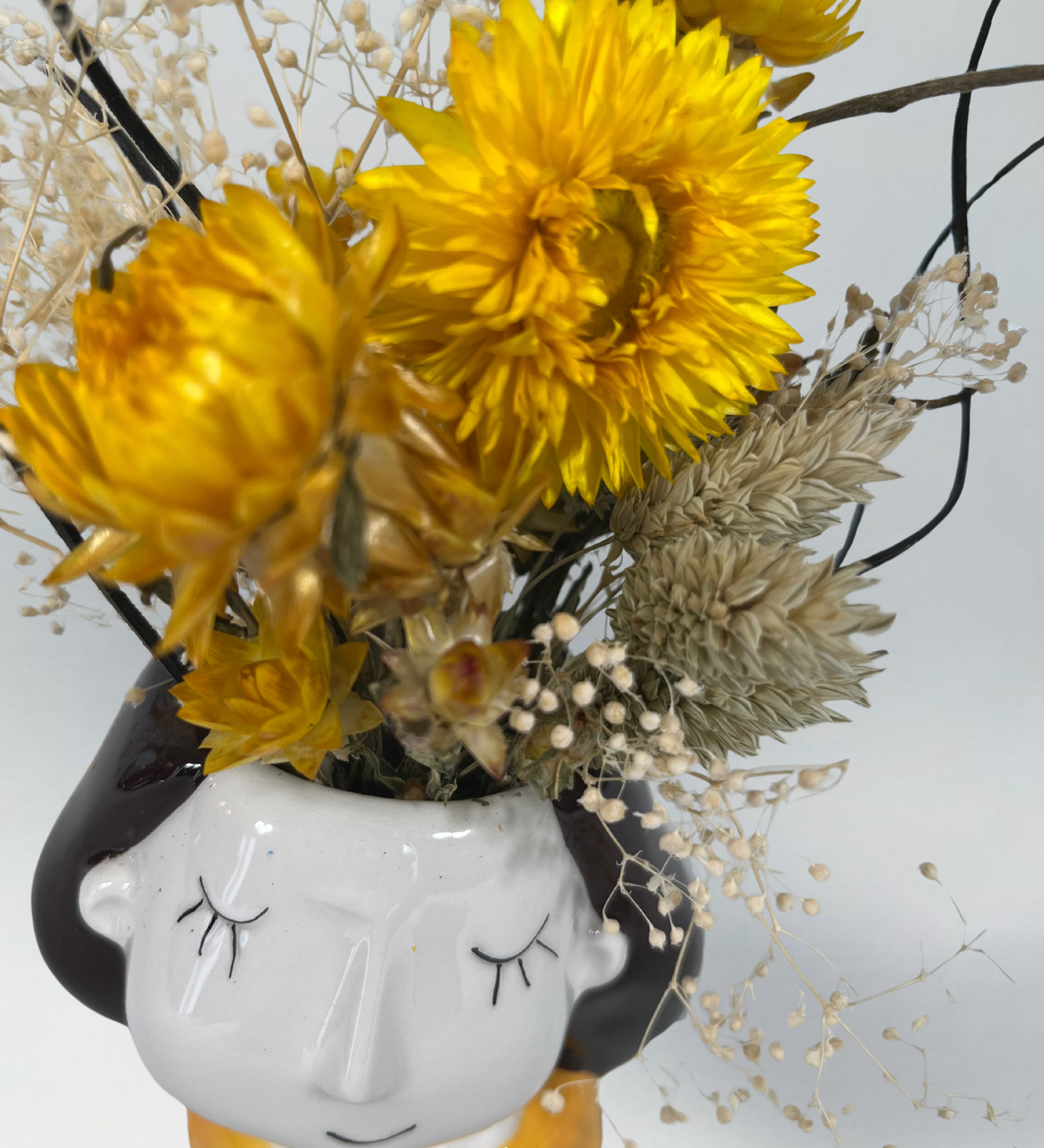 Vase shaped like a face with yellow flowers and dried elements on a white background