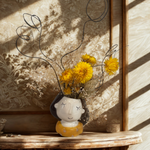 Decorative vase with dried flowers on a wooden shelf against floral wallpaper.