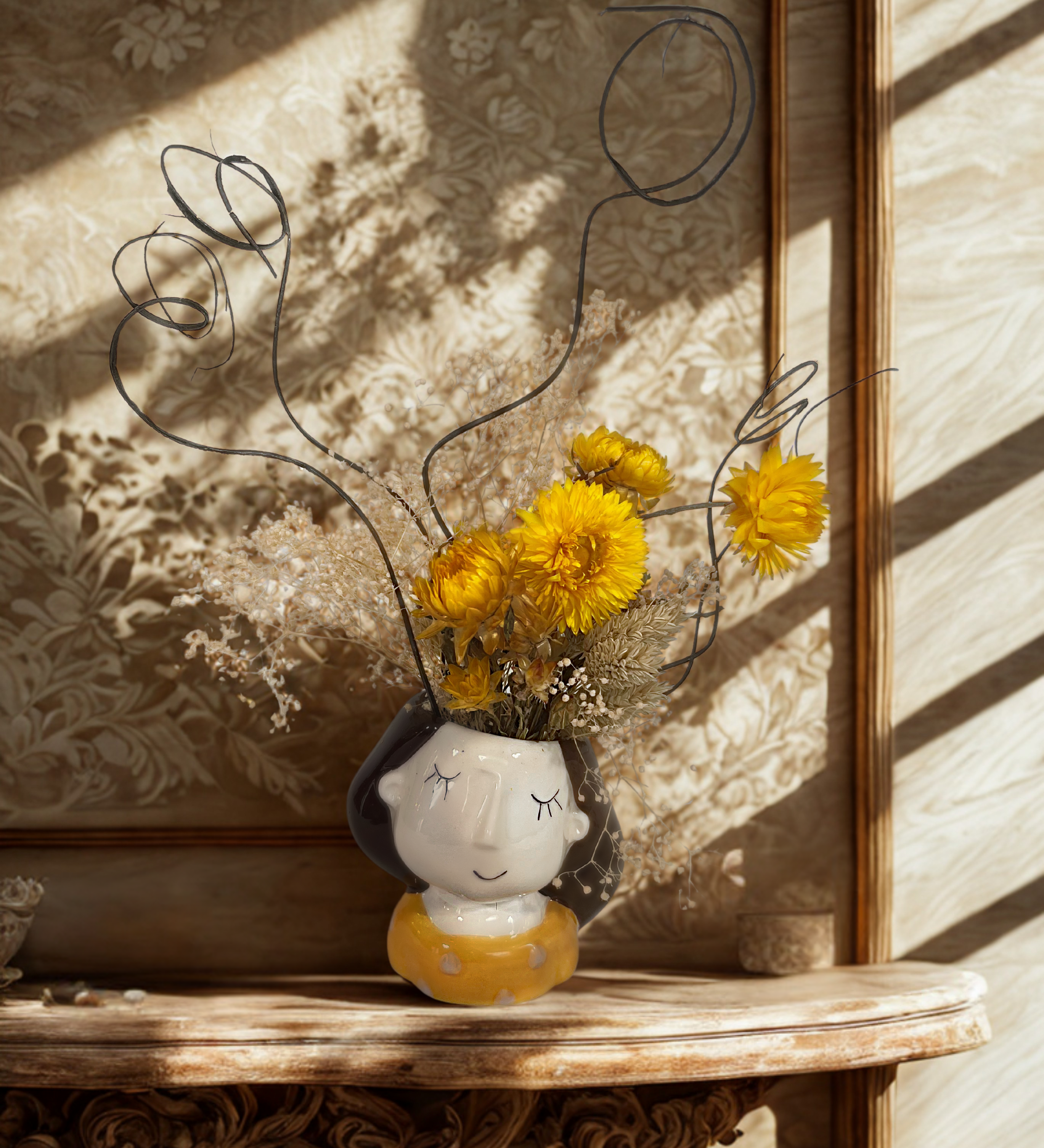 Decorative vase with dried flowers on a wooden shelf against floral wallpaper.