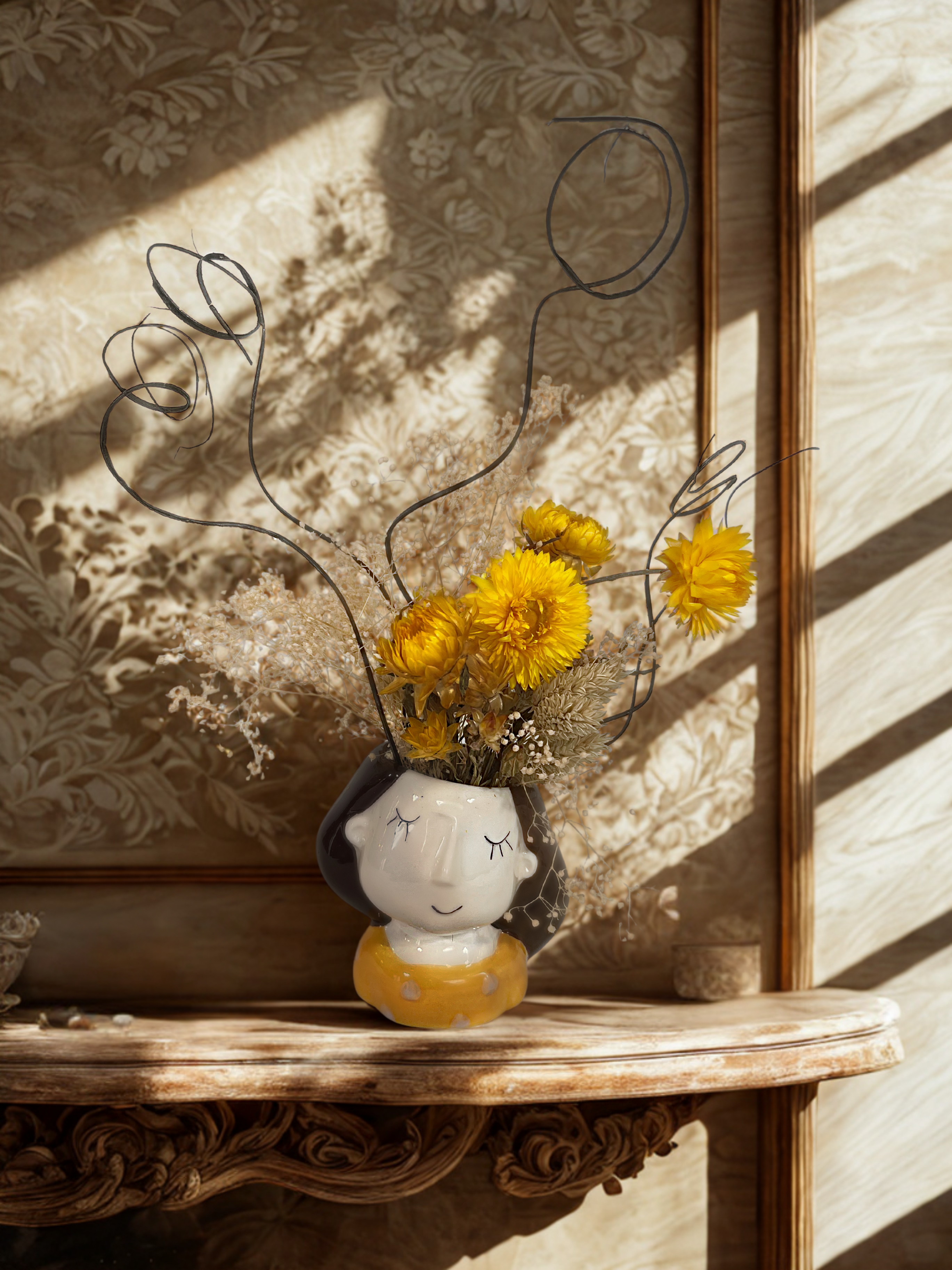Decorative vase with dried flowers on a wooden shelf against floral wallpaper.