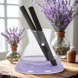 Glass decanter with black reeds on a wooden surface with lavender flowers in the background