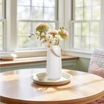 White ceramic vase with matte finish and faux leather handle holding flowers on a table in a bright room.