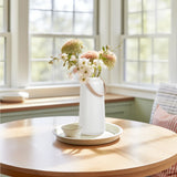 White ceramic vase with matte finish and faux leather handle holding flowers on a table in a bright room.