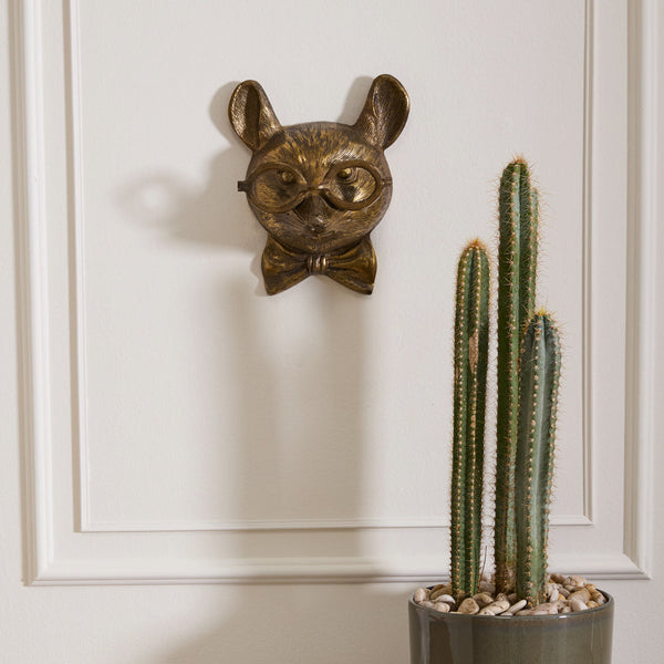 Decorative wall mask with a bow tie above a potted cactus on a white paneled wall.
