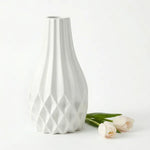 A white ceramic vase with a flared neck and geometric diamond-patterned base, shown on a clean white background with three pale tulips beside it.