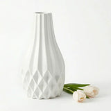 A white ceramic vase with a flared neck and geometric diamond-patterned base, shown on a clean white background with three pale tulips beside it.