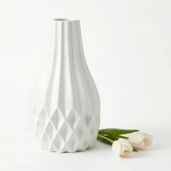 A white ceramic vase with a flared neck and geometric diamond-patterned base, shown on a clean white background with three pale tulips beside it.