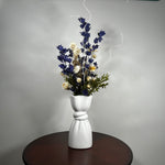 A white ceramic vase with a sculptural, twisted base holding a dried floral arrangement of blue, cream, and green flowers, set on a dark round table against a gray wall.