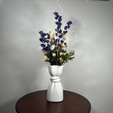 A white ceramic vase with a sculptural, twisted base holding a dried floral arrangement of blue, cream, and green flowers, set on a dark round table against a gray wall.
