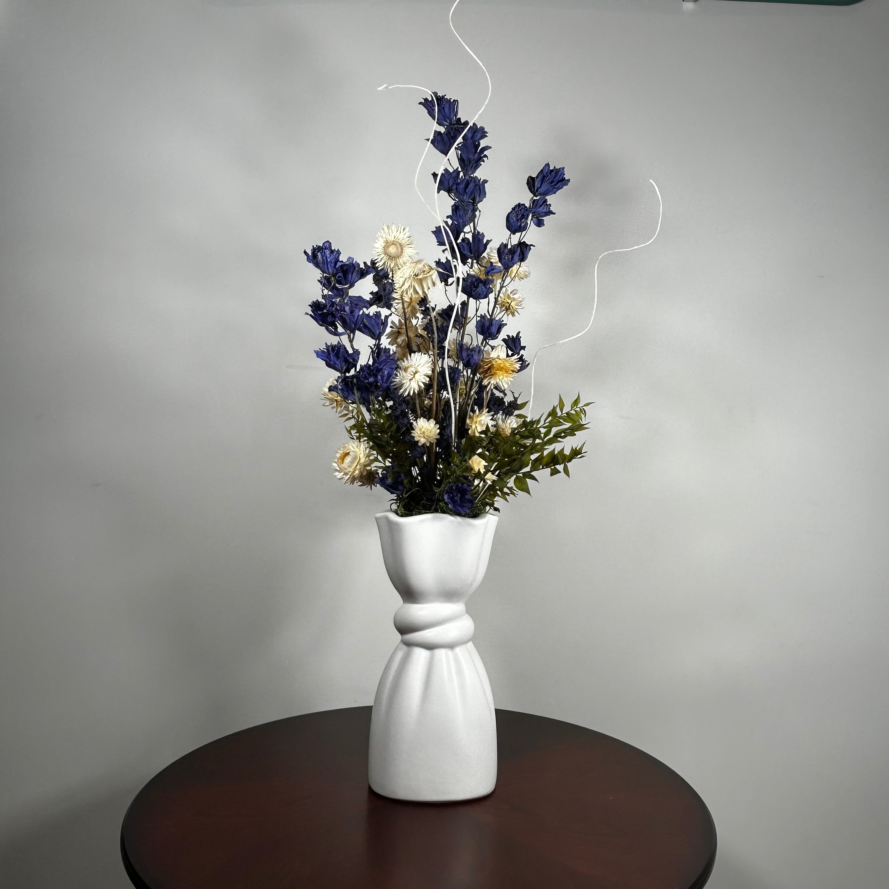 A white ceramic vase with a sculptural, twisted base holding a dried floral arrangement of blue, cream, and green flowers, set on a dark round table against a gray wall.