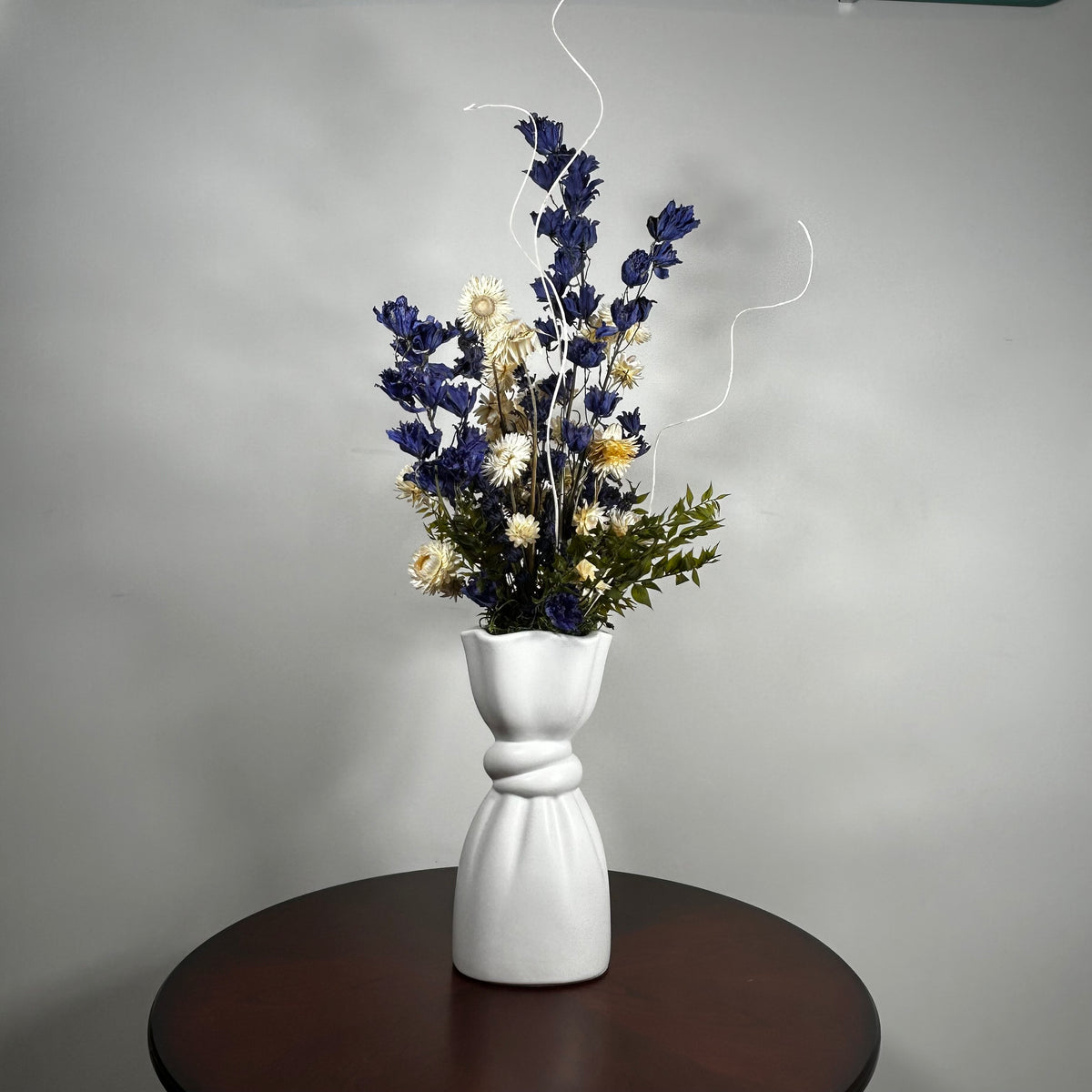 A white ceramic vase with a sculptural, twisted base holding a dried floral arrangement of blue, cream, and green flowers, set on a dark round table against a gray wall.