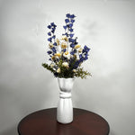 A white ceramic vase with a sculptural, face-like neck and flared rim, holding a dried floral arrangement of blue delphiniums, cream daisies, and green foliage, set on a dark round wooden table against a light gray wall.