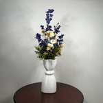 A white ceramic vase with a sculptural, flared top and ribbed base, holding a floral arrangement of blue delphiniums, cream daisies, and green foliage, set on a dark round wooden table against a light gray wall.
