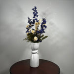A white ceramic vase with a sculptural, flared base and a twisted center detail, filled with a dried floral arrangement featuring dark blue, cream, and green elements, set on a dark round wooden table against a neutral gray wall.