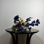 A dried floral arrangement in deep blue, cream, and olive green tones, displayed on a dark round table against a plain light gray wall.