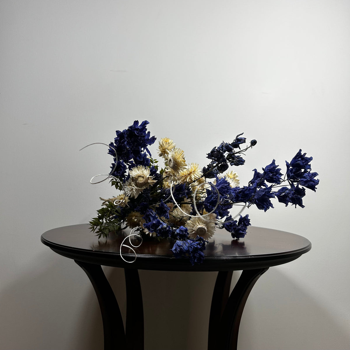 A dried floral arrangement in deep blue, cream, and olive green tones, displayed on a dark round table against a plain light gray wall.