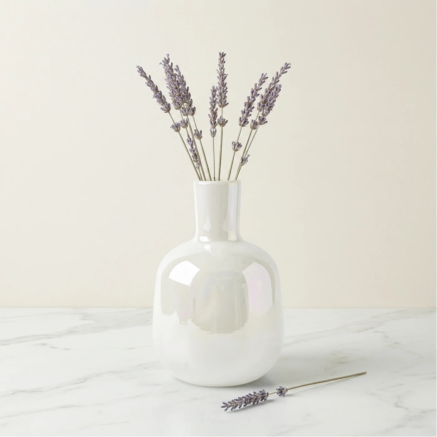 Mini bud vase features a sleek bottle shape and a rainbow lustre glass finish that catches the light beautifully. Shown here with sprigs of lavender on a white marble table with a white background.
