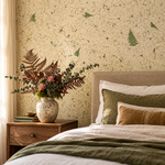 A styled bedroom corner featuring Stone Pine Forest wallpaper with a speckled cream background and scattered green fern motifs. A wooden nightstand holds a ceramic vase filled with dried ferns and greenery, beside a bed with cream bedding, olive green pillows, and a tan accent pillow. 3