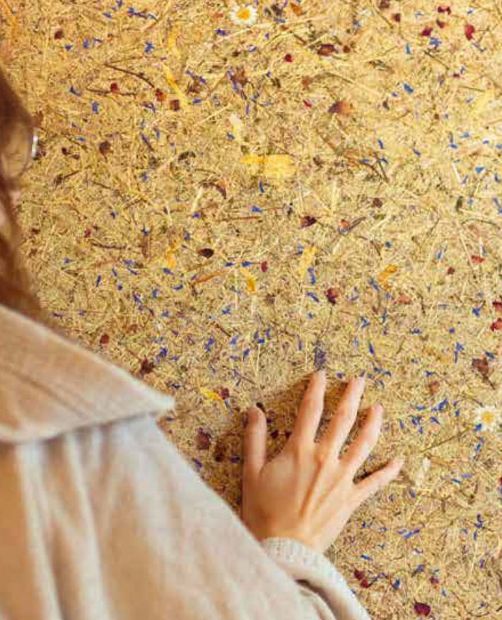 A close-up view of a person's hand touching a textured cork floor with embedded dried flowers and small colorful flecks, with part of their beige coat visible in the foreground.