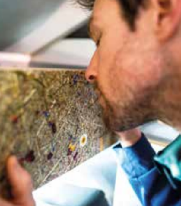 A close-up lifestyle shot of a man smelling a textured, earthy-toned sample of dried botanical fiber or felt material held in his hand.