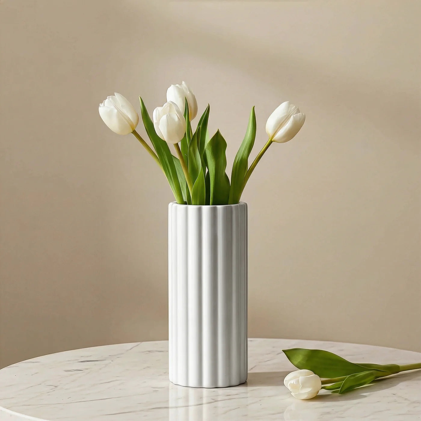 A tall cylindrical vase, whose silhouette is softened by delicate vertical ribbing, creating subtle movement as light shifts across its glossy white surface. Shown here with white tulips on a marble table with a beige background.