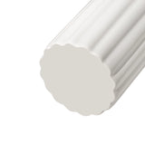 A close-up of the bottom of a tall cylindrical vase, whose silhouette is softened by delicate vertical ribbing, creating subtle movement as light shifts across its glossy white surface. 