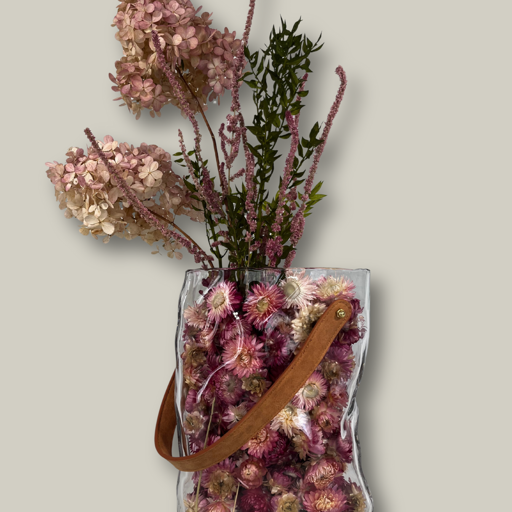 Clear glass vase shaped like a bag filled with dried flowers on a light gray background