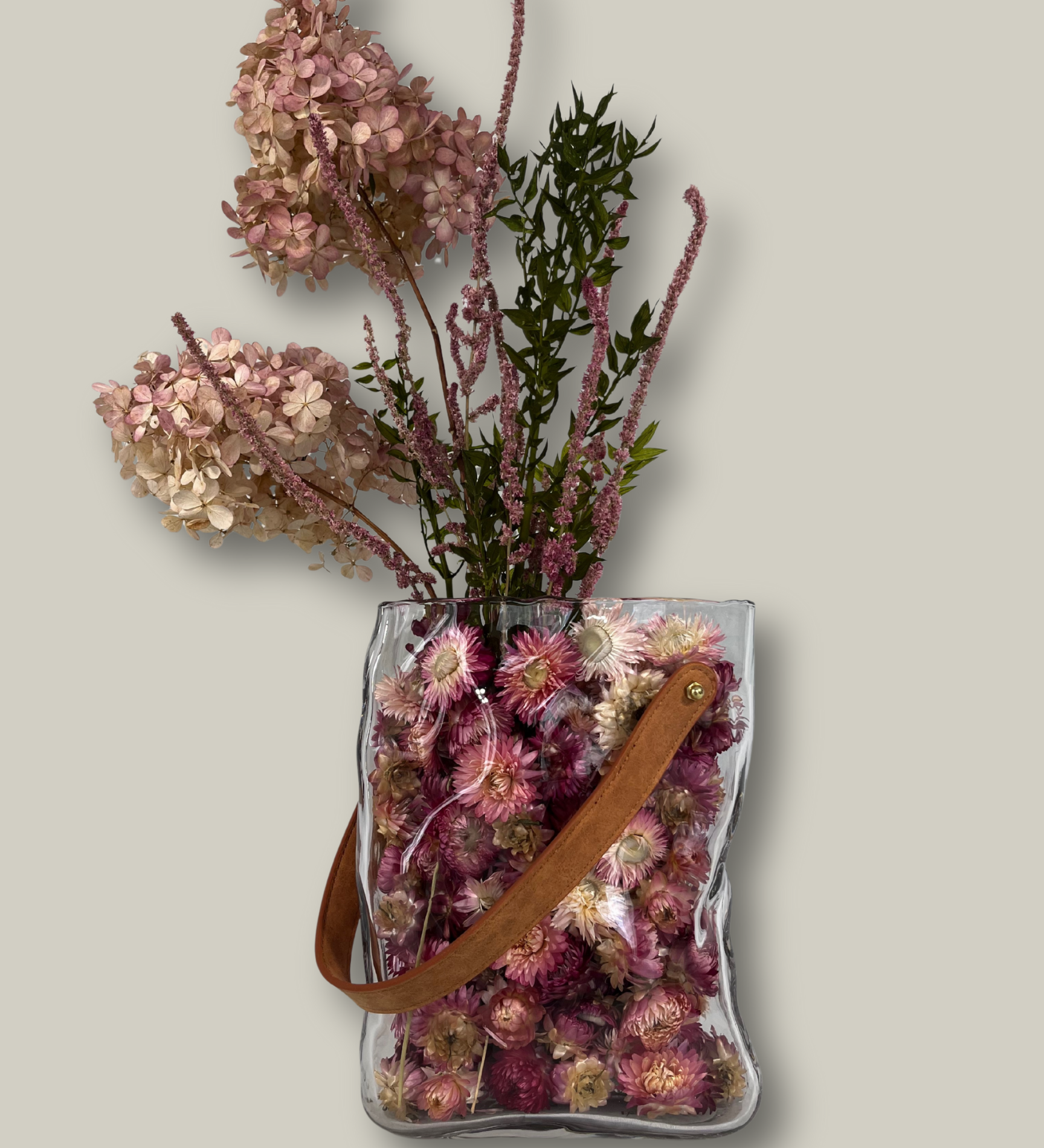Clear glass vase shaped like a bag filled with dried flowers on a light gray background