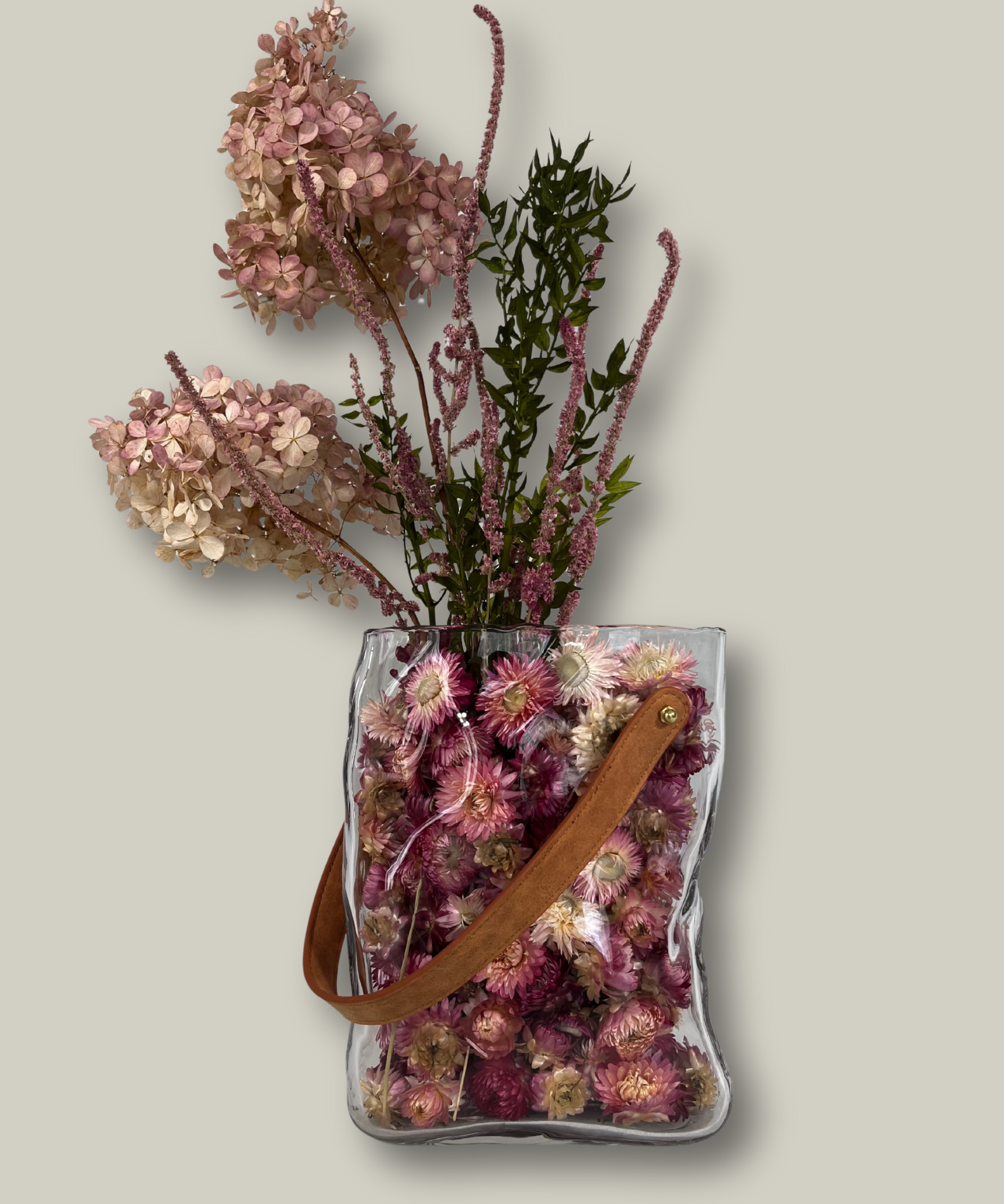 Clear glass vase shaped like a bag filled with dried flowers on a light gray background