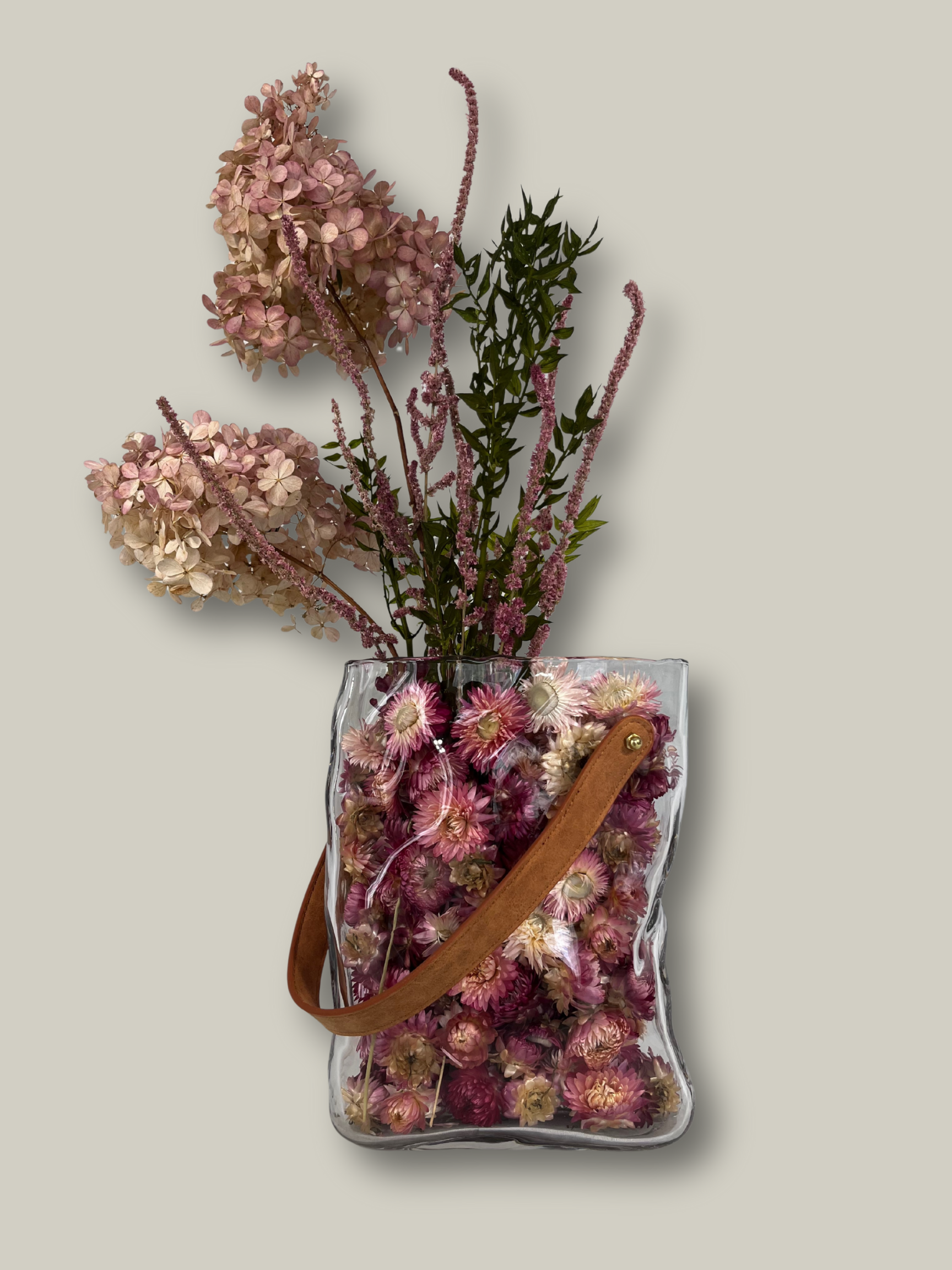 Clear glass vase shaped like a bag filled with dried flowers on a light gray background