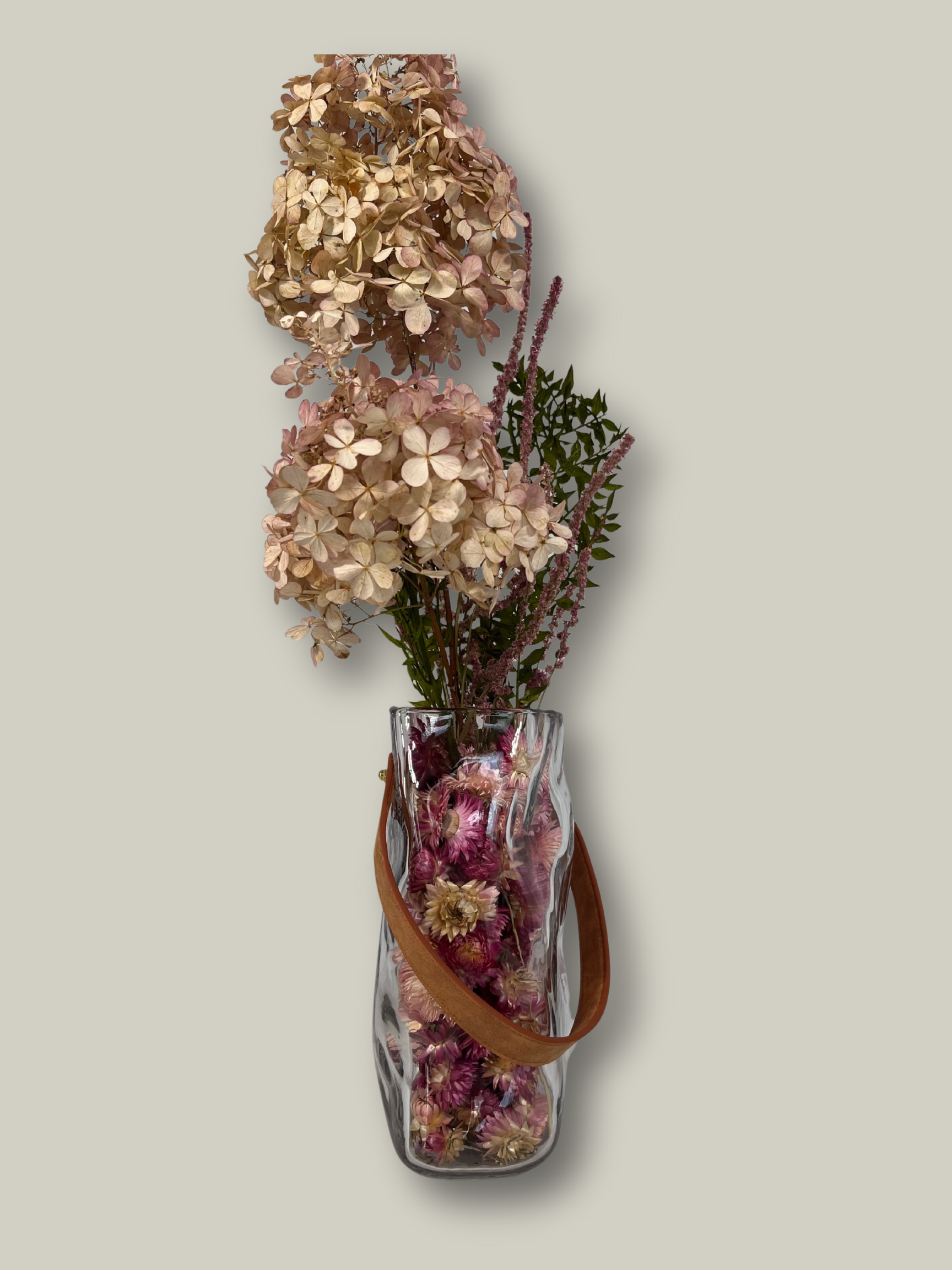 Clear glass vase shaped like a bag with dried flowers on a light gray background