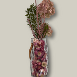 Clear glass vase shaped like a bag with pink dried lowers on a light gray background