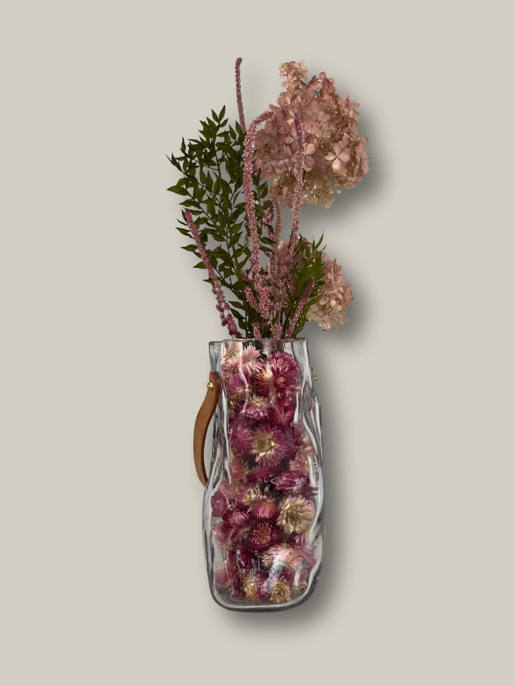 Clear glass vase shaped like a bag with pink dried lowers on a light gray background