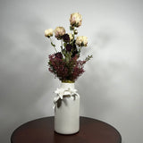 A white ceramic vase with sculpted white flowers at the neck, holding a dried floral arrangement with cream, burgundy, and dark purple blooms, displayed on a dark round table against a light gray wall.