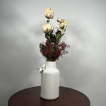 A white ceramic vase with a textured finish, decorated with a small white bow, holding a dried flower arrangement with cream and burgundy tones, set on a dark round table against a light gray wall.