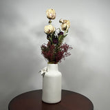A white ceramic vase with a textured finish, decorated with a small white bow, holding a dried flower arrangement with cream and burgundy tones, set on a dark round table against a light gray wall.